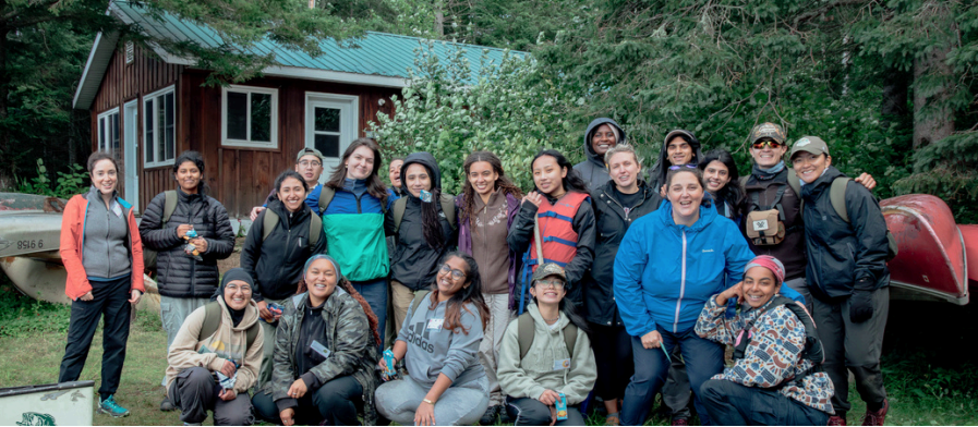 A group of people standing outside, with a cabin, trees, and canoes behind them. 